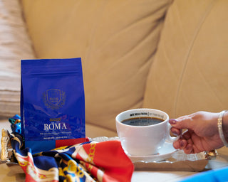 Hand holding a cup of espresso beside a Parisi Roma coffee bag on a cozy table setting with warm tones