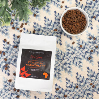 A white bowl of Apex Ethiopian Yirgacheffe coffee beans rests on a blue patterned cloth beside a green plant.