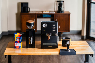 Wide shot of the Eureka Mignon Zero 65 espresso grinder on a wooden table with a coffee bag and LUCCA machine in the background