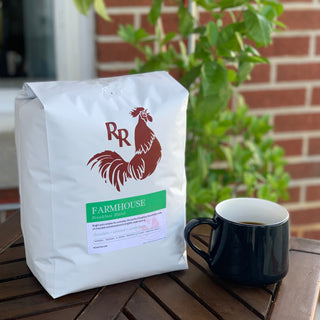 White coffee bag with a red rooster logo and green Farmhouse label on a wooden table beside a black mug