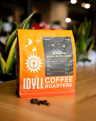 Glow in the Dark coffee bag with a dark label illustration on orange packaging placed on a wooden table with coffee beans nearby