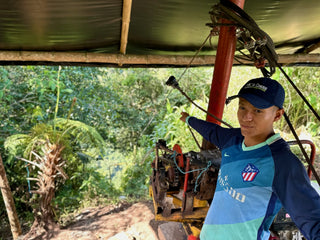 Farm worker in a blue shirt and cap operating processing equipment beneath a shade canopy on a Colombia coffee farm