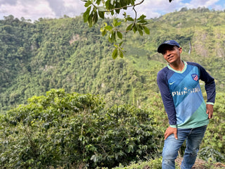 Smiling farmer in a blue cap and light blue shirt among coffee plants on a hillside