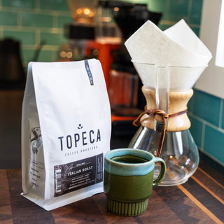 Topeca Italian Roast coffee bag in the foreground with a glass Chemex and a green mug on a wooden counter in a cafe setting