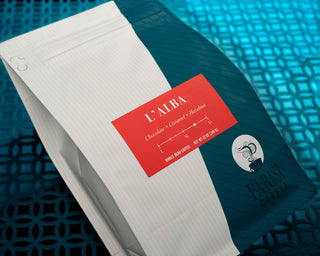 L Alba coffee bag with red label lying on white lined paper atop a teal surface