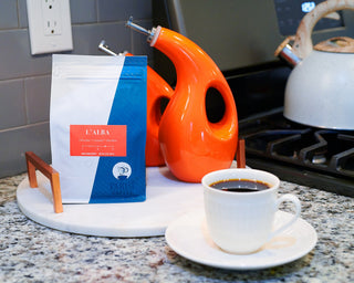 Parisi L Alba coffee bag on a marble kitchen counter with an orange kettle and a cup of coffee nearby