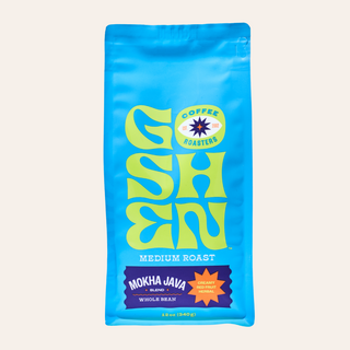 Close up of a bright blue Mokha Java coffee bag with a bold yellow logo and a cup of coffee in the foreground.