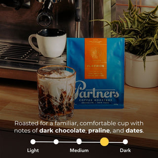 Glass of iced coffee beside a blue Flatiron espresso bag on a wooden counter.