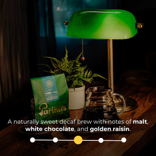 Partners Ghost Town Decaf bag on a wooden surface beside a green glass lamp and plant.