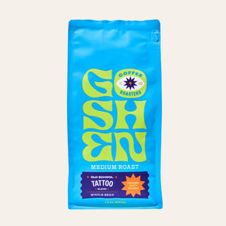 Blue Old School Tattoo whole bean coffee bag with medium roast label beside a cup of coffee on a colorful table