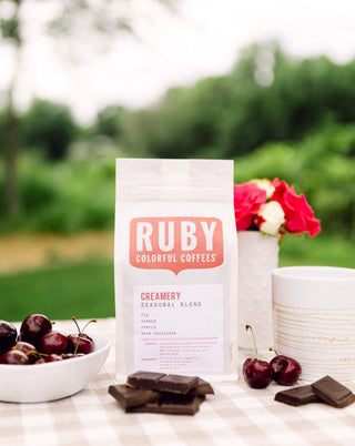 Creamery Seasonal Blend coffee bag with Ruby Colorful Coffees logo outdoors beside cherries and chocolate on a checkered table.