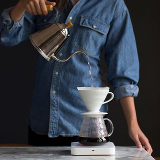 Barista in a denim shirt pours hot water over a pour-over setup with a white dripper and glass carafe.