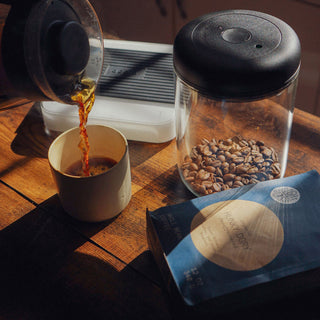 Brewing Hunky Dory Decaf coffee a stream pours into a mug beside the blue decaf bag and a jar of beans on a wooden table