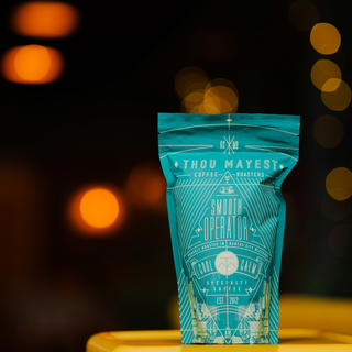 Teal Smooth Operator coffee bag on a yellow table with warm orange bokeh lights in the background