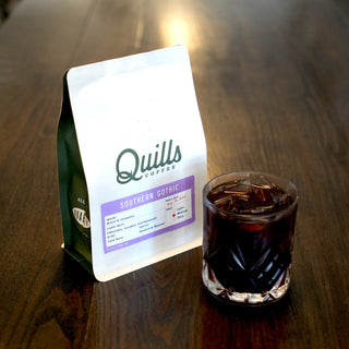 Quills Southern Gothic Cold Brew bag on a wooden table with a glass of iced coffee beside it