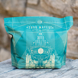 Teal Thou Mayest coffee bag on a wooden surface with logo and cityscape graphic