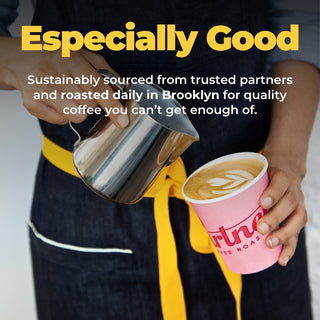 Barista pours hot coffee from a stainless kettle into a pink latte cup while wearing a dark apron with a yellow belt.