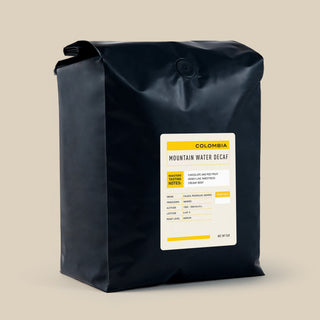 Close up of a black coffee bag with Mountain Water Decaf Colombia label on a beige backdrop