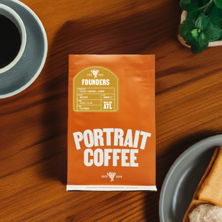 Orange Portrait Coffee bag on a wooden table with a cup of coffee and a pastry nearby