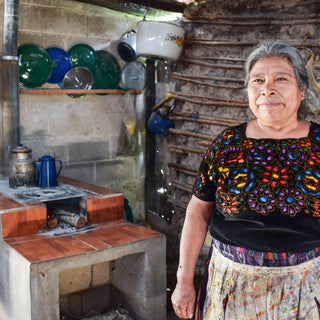 Guatemalan woman in an embroidered blouse stands in a rustic kitchen beside a clay stove