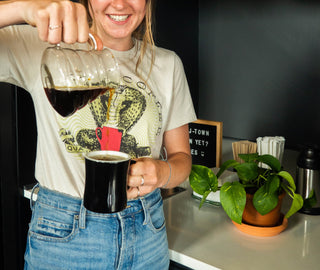 Smiling person in a Quills Coffee shirt pours coffee from a glass carafe into a cup beside a Quills bag