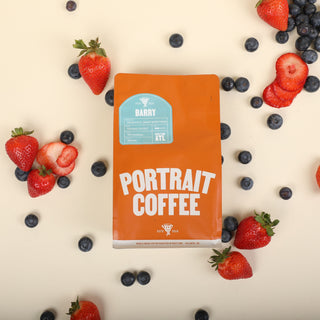 Overhead shot of an orange Barry coffee bag surrounded by strawberries and blueberries on a pale beige surface.