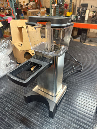 Open box Moccamaster KBGT polished silver coffee grinder with clear hopper on a workbench.