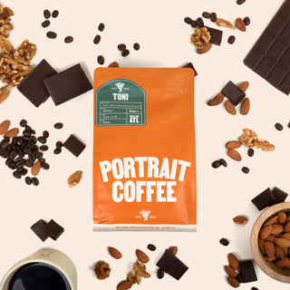 TONI Portrait Coffee bag surrounded by chocolate chunks almonds walnuts and scattered coffee beans on a beige surface with a cup of coffee nearby