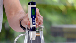Handheld water tester held over a glass coffee carafe during testing